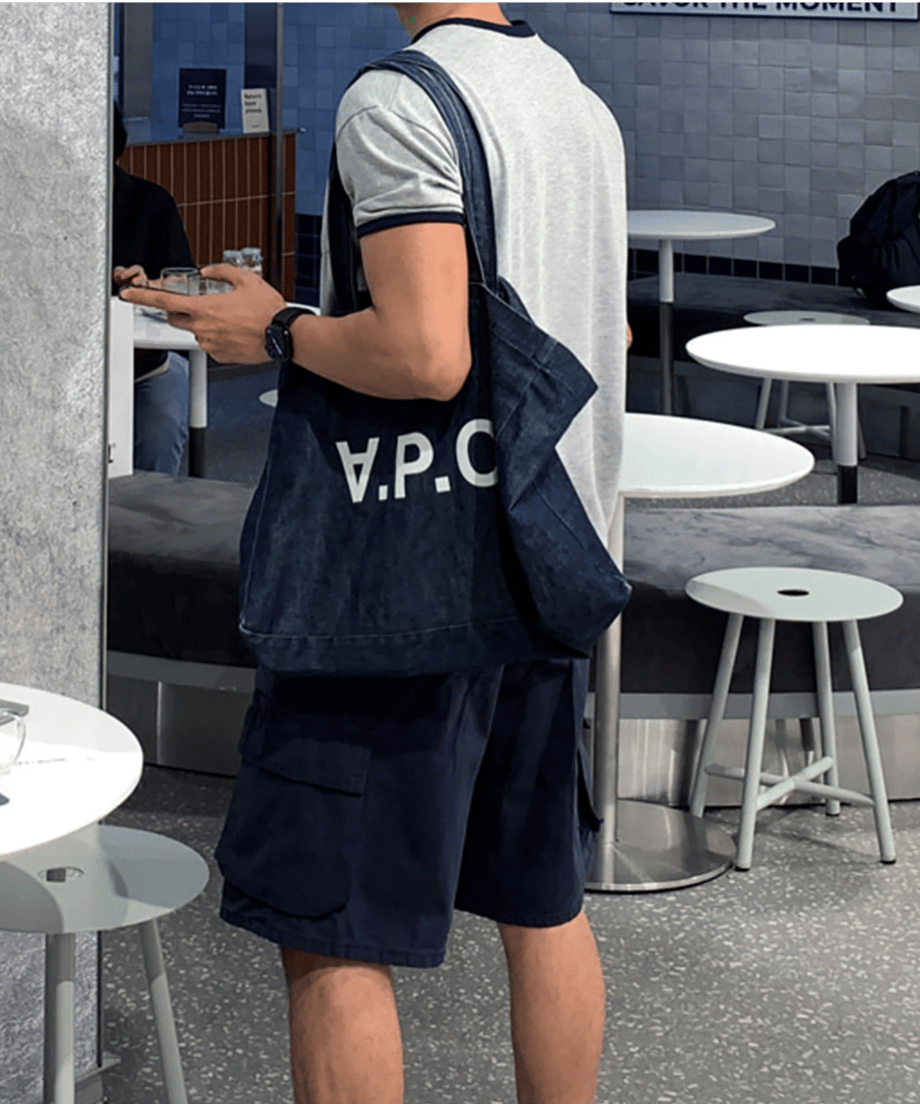 リンガーTシャツにネイビーのショーツを合わせ、ロゴ入りデニムバッグを肩掛けした男性の、カフェ店内での斜め後ろからの全身スタイリング。
