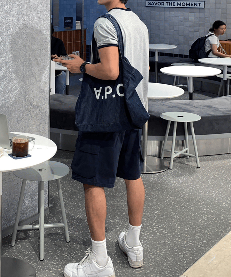リンガーTシャツにネイビーのショーツ、白ソックス、白スニーカーを合わせ、ロゴ入りデニムバッグを肩掛けした男性の、カフェ店内での斜め後ろからの全身スタイリング。