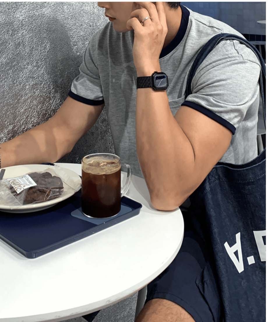 リンガーTシャツを着用し、カフェの丸テーブルでアイスコーヒーとクッキーを前に、スマートウォッチをはめた腕を机につけてリラックスする男性のクローズアップ。
