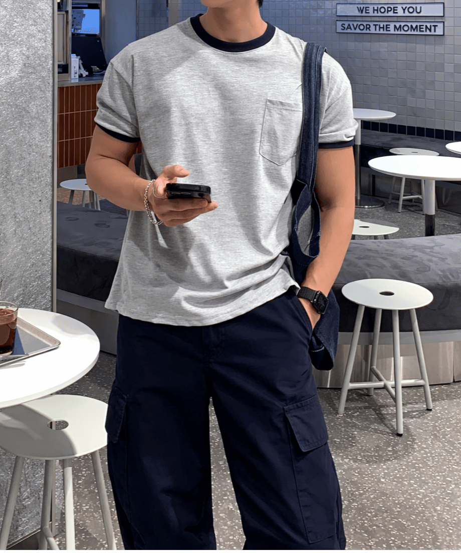 左胸のポケットがアクセントのリンガーTシャツに、ネイビーのワイドカーゴパンツを合わせた、カフェの店内を背景にした男性のスタイリッシュな立ち姿。