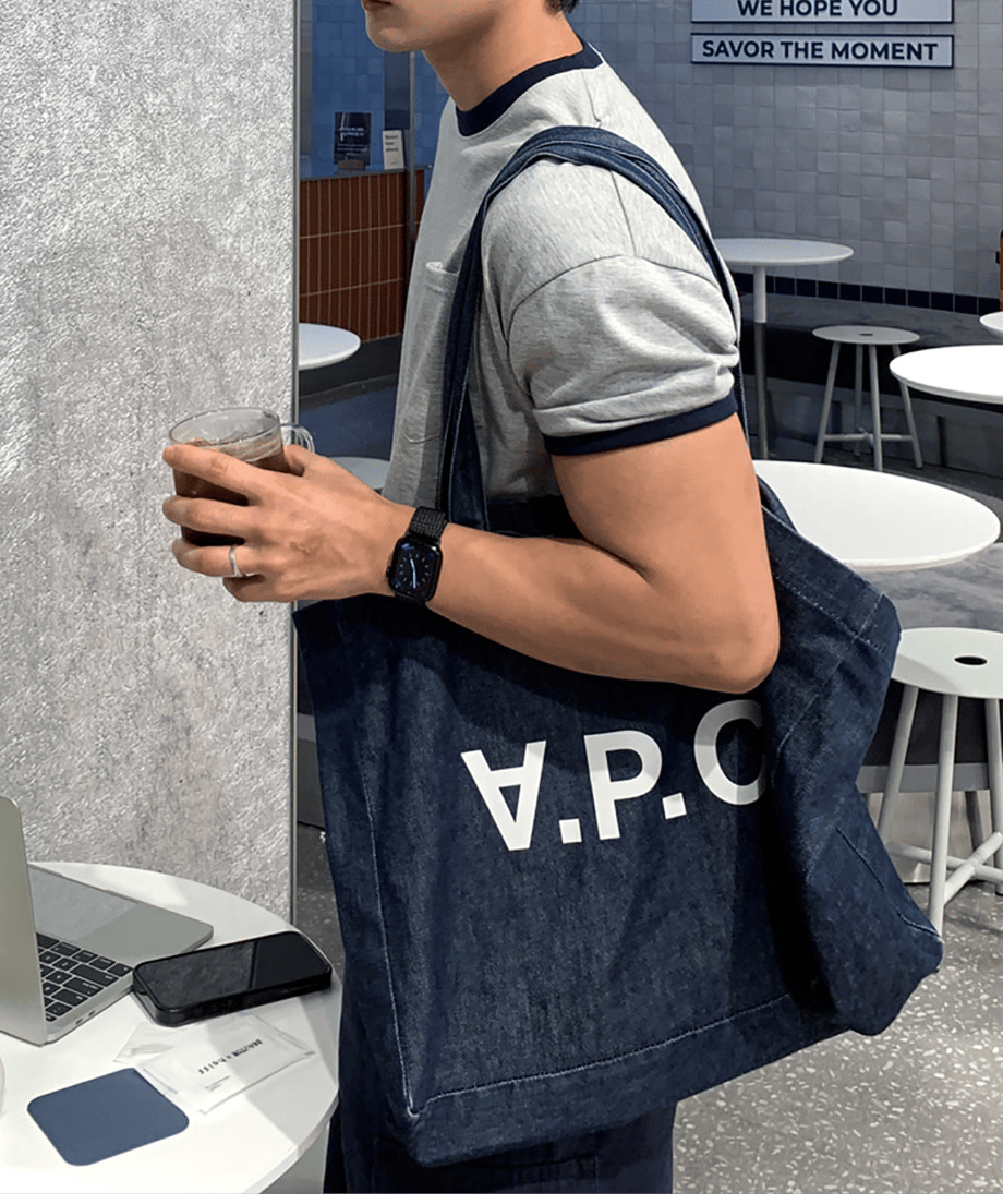 リンガーTシャツを着用し、ロゴ入りのデニムトートバッグを肩に掛けながら、カフェのテーブル席でアイスコーヒーを持つ男性のサイドショット。