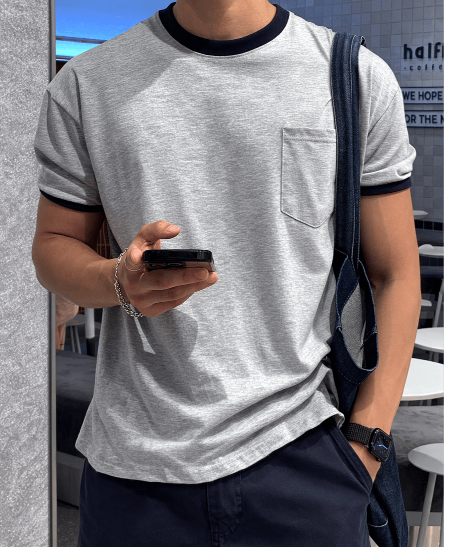 ライトグレーにネイビーのリブが映えるリンガーポケットTシャツを着用し、片手をポケットに入れながらスマートフォンを操作する男性のフロントカット。