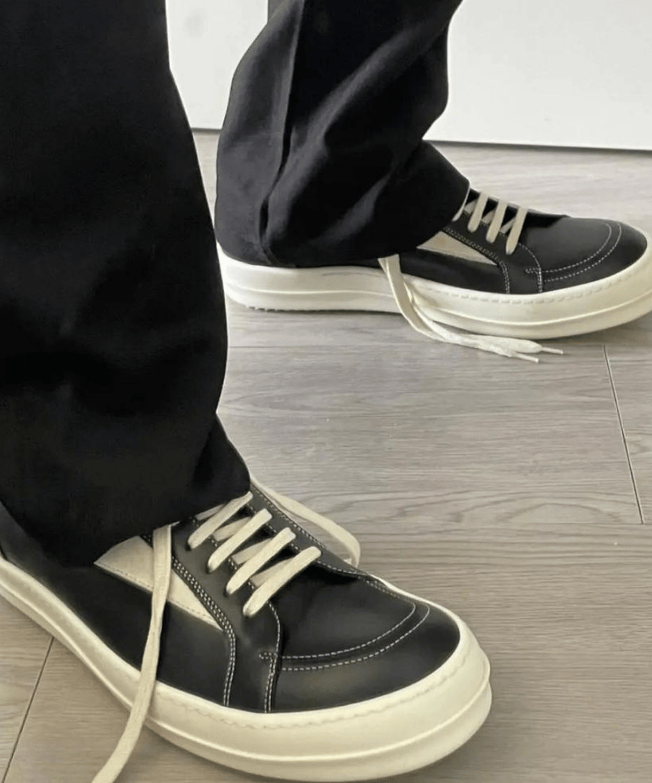 黒いパンツの裾から覗く、オフホワイトの靴紐が結ばれた黒いレザースニーカーをフローリングの床の上で近くから撮影した写真。