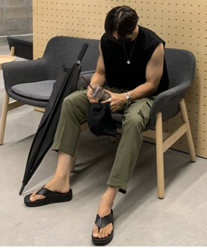 ブラックのマッスルタンクトップにカーキのカーゴパンツ、黒のトングサンダルを合わせ、黒い傘を横に置いてソファに深く座り込む男性の全身姿。