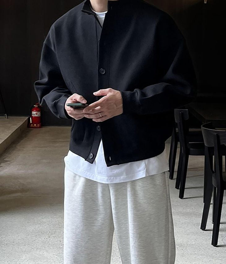 ブラックのスタンドカラーカーディガンと白いインナー、ライトグレーのパンツを着用し、スマートフォンを両手で持つ男性の正面姿。