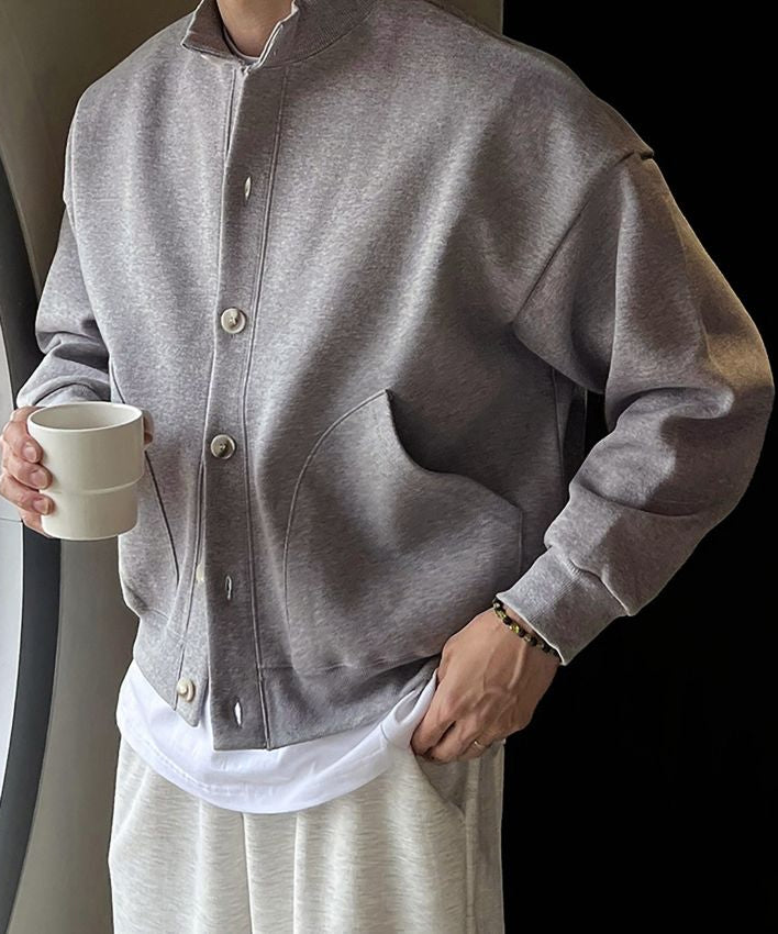 グレーのスタンドカラーカーディガンを着用し、片手をポケットに入れて白いマグカップを持つ男性の前身頃アップ姿。