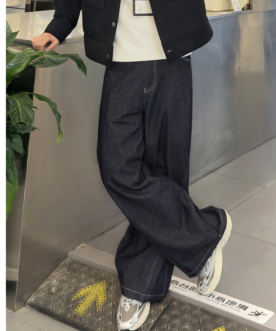 足を軽く交差させ、立体的なシルエットのワイドバルーンデニムパンツを穿いて立つ男性の屋内のクローズアップ写真。