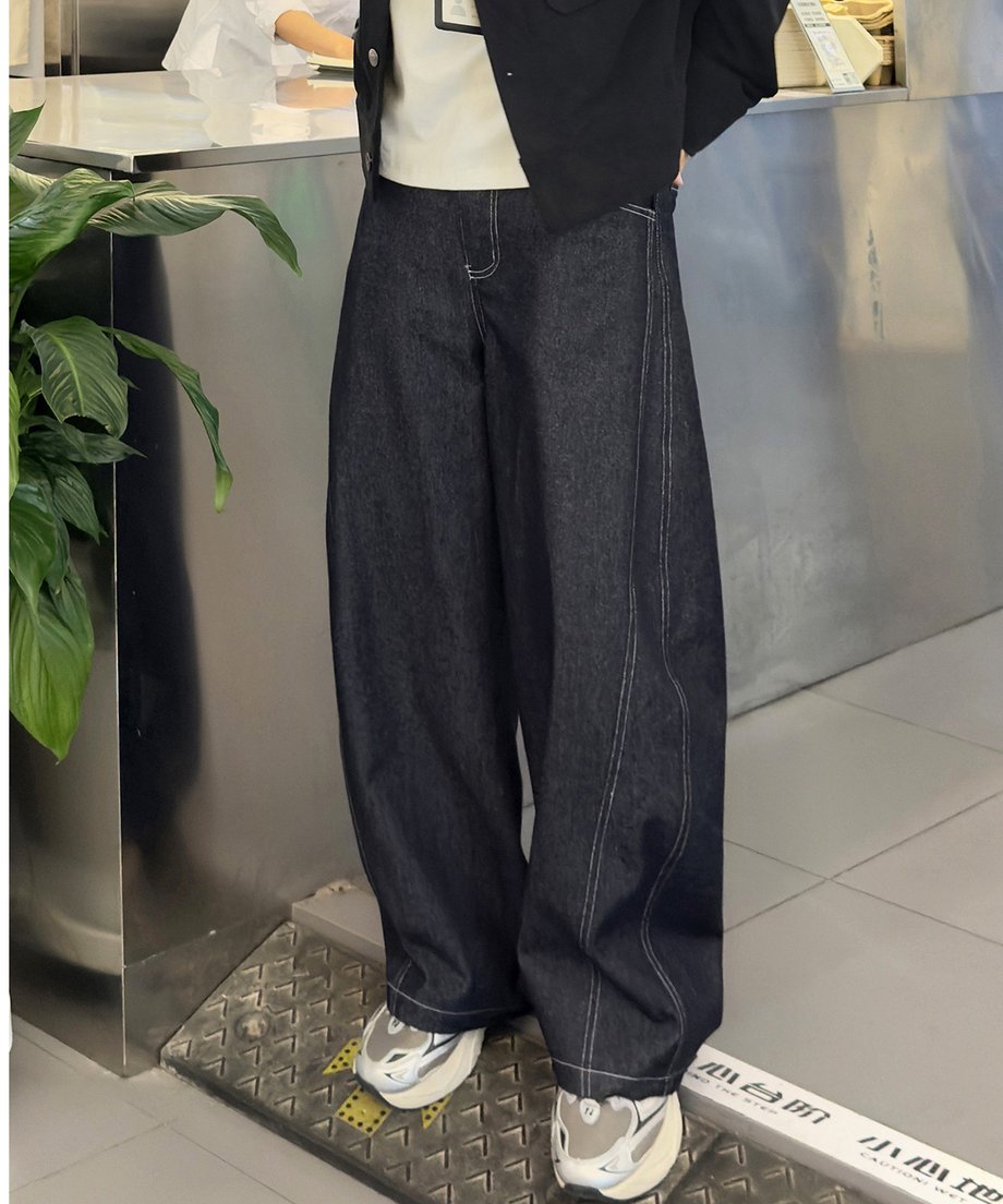 メタリックなカウンターの横に立ち、白ステッチが施されたダークネイビーのワイドバルーンデニムパンツにグレーのスニーカーを合わせた男性の写真。