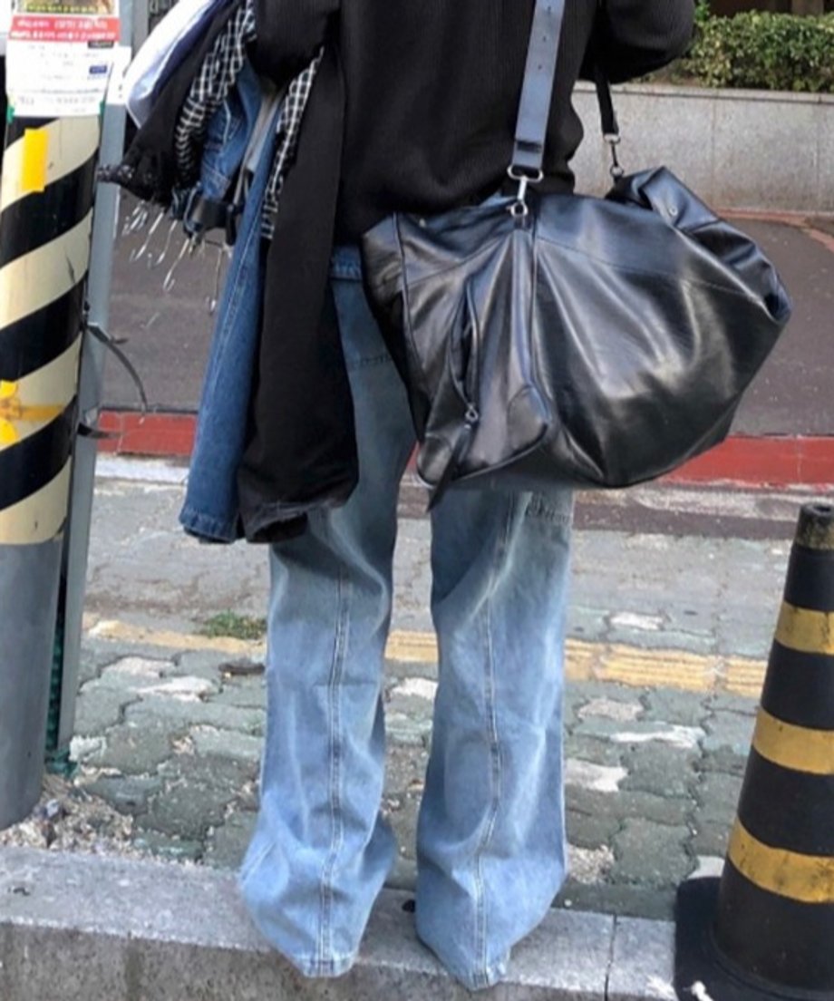 黒のカットソーにブルーのバギーデニムカーゴパンツを穿き、黒の大きなレザーバッグを背負って歩道に立つ男性の後ろ姿。
