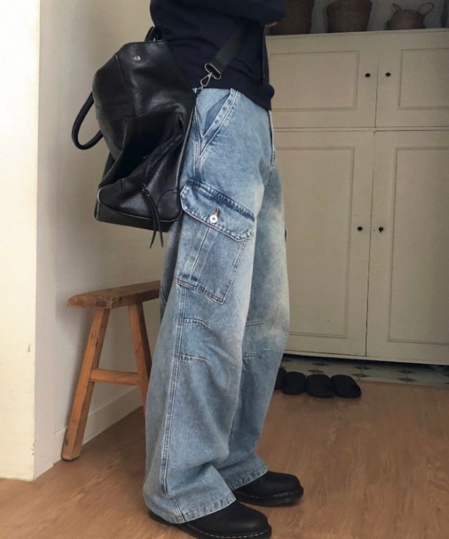 室内で黒の大きなレザーバッグを肩に掛け、大きなフラップポケット付きのブルーワイドデニムカーゴパンツを穿いた男性の横向きのカット。