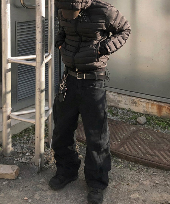 屋外のメタルラックの横で、黒のダウンジャケットとフードを被り、立体裁断のブラックユーティリティバギーパンツを穿いて佇む男性の全身スタイリング。