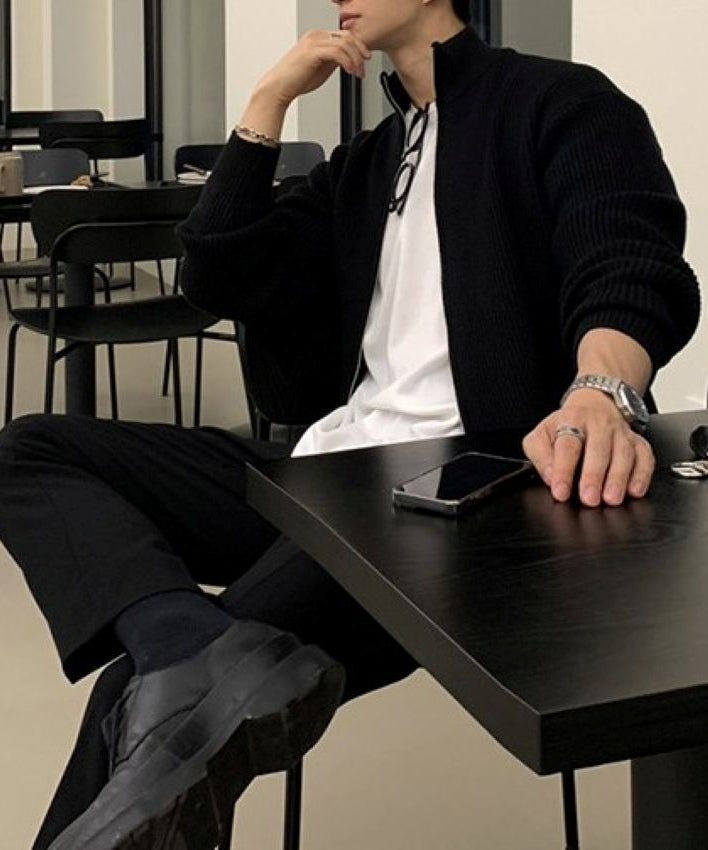 ブラックのジップアップニットカーディガンに白Tシャツ、黒のパンツ、黒の革靴を合わせ、カフェの椅子に腰掛ける男性の全身コーディネート姿。