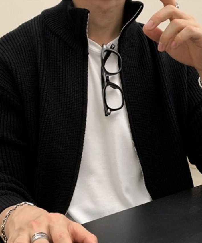 ブラックのジップアップニットカーディガンを着用し、カフェの黒いテーブル席につく男性のバストアップ姿。胸元にサングラスを掛けたスタイリング。