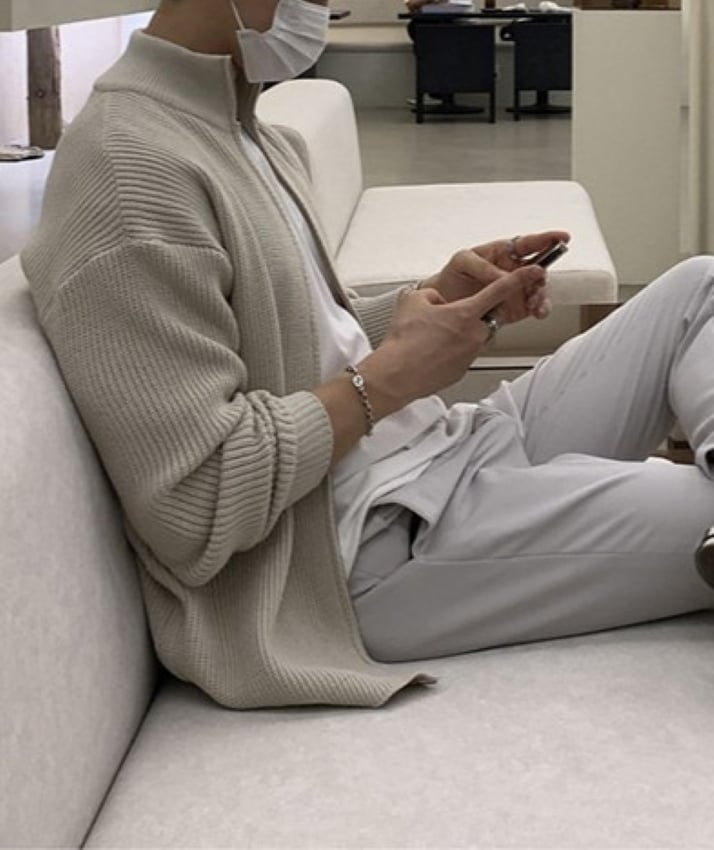 ベージュのジップアップニットカーディガンを着用し、白いソファに深く腰掛けてスマートフォンを操作する男性の横姿。