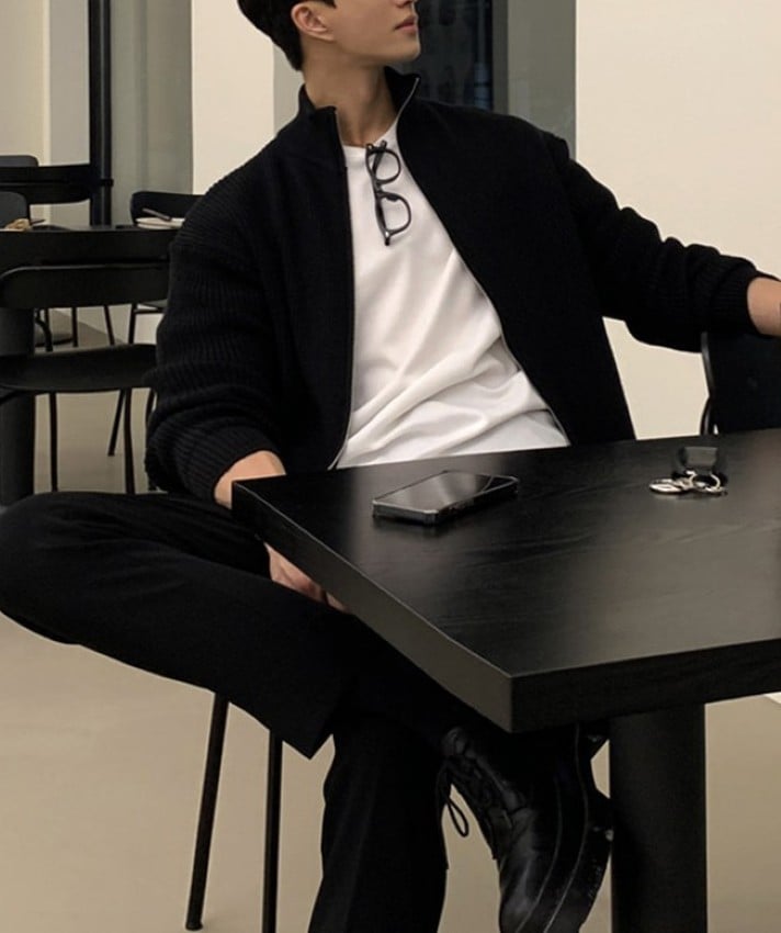 ブラックのジップアップニットカーディガンを着用し、カフェの椅子に座って外を見つめる男性のスタイリングカット。