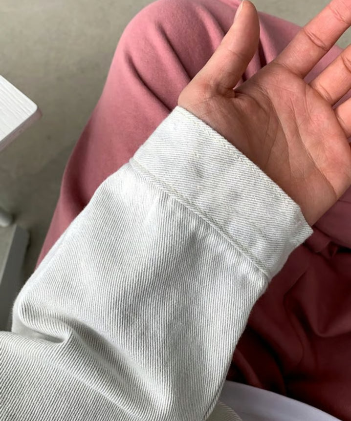 ホワイトのバンドカラーデニムジャケットを着用し、くすみピンクのパンツを背景に、手元と袖口のステッチやカフス部分をクローズアップしたカット。