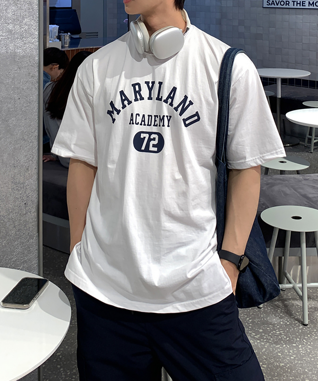 ホワイトのメリーランドアカデミーTシャツを着用し、首元に白いヘッドホンを掛け、ネイビーのパンツのポケットに手を入れた男性のアップ姿。