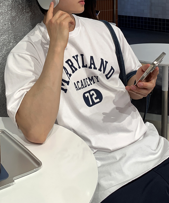 ホワイトのメリーランドアカデミーTシャツを着用してカフェの白いテーブル席につき、スマートフォンを持ちながらヘッドホンを耳に当てる男性の上半身姿。