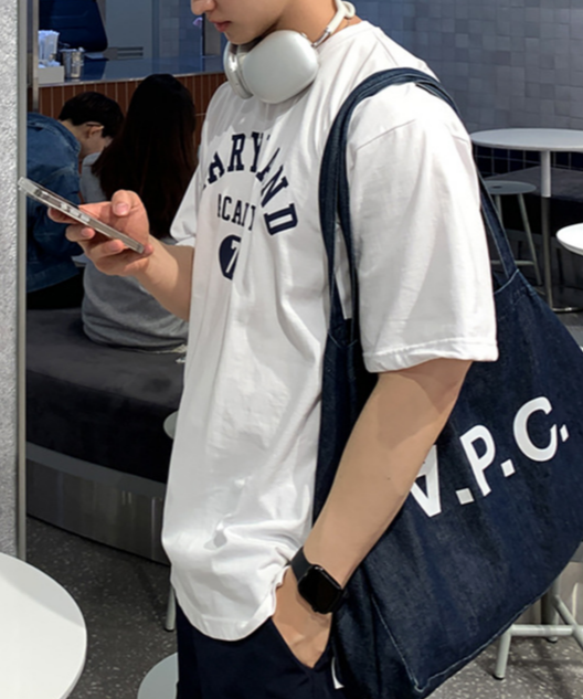 ホワイトのメリーランドアカデミーTシャツを着用し、デニムの大型トートバッグを肩に掛けながらスマートフォンを操作する男性の横からのシルエット。