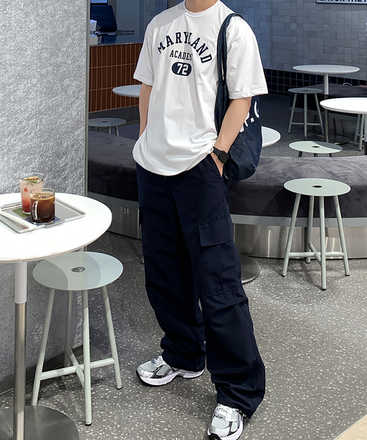 ホワイトのメリーランドアカデミーTシャツにネイビーのカーゴパンツを合わせ、デニムのトートバッグを肩に掛けて立つ男性の全体バランス。