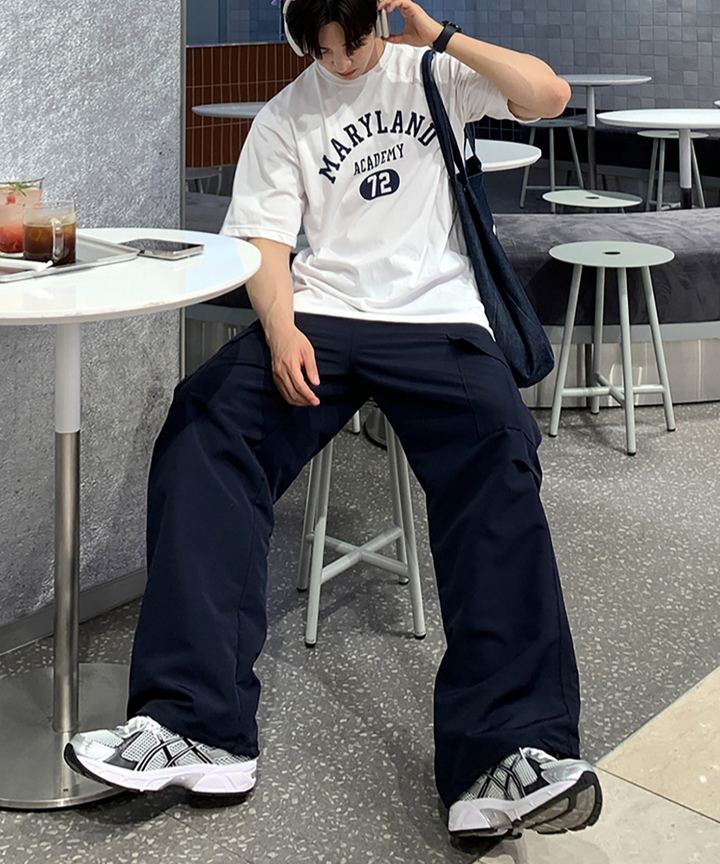 ホワイトのメリーランドアカデミーTシャツにネイビーのワイドカーゴパンツ、白スニーカーを合わせ、カフェの椅子に座ってヘッドホンに手を添える男性の全身コーディネート。