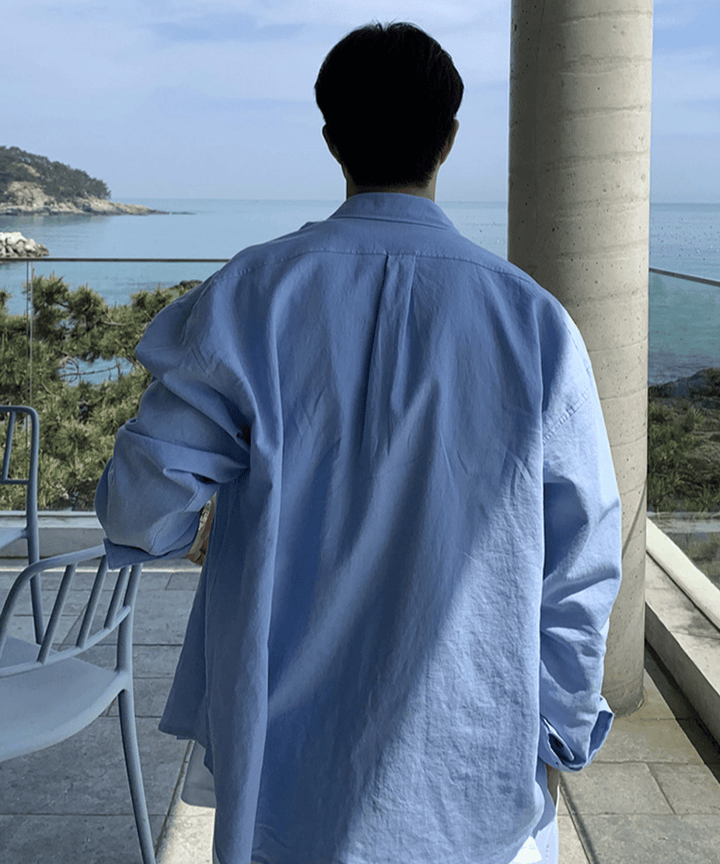 ゆったりとしたスカイブルーのリネンシャツを着用した男性の、海をバックにした開放感あふれるバックシルエット。
