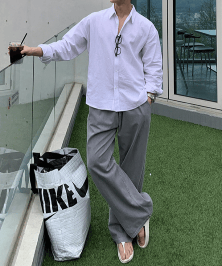 ホワイトのカジュアルシャツにグレーのルーズパンツ、足元に白のトングサンダルを合わせ、大きなロゴ入りトートバッグを足元に置いて立つ男性の全身スタイリング。