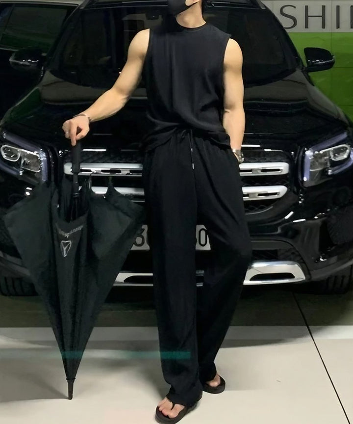 ブラックのプリーツノースリーブセットアップにサンダルを合わせ、黒い傘を右手に持って車の前に佇む男性の全身フロントショット。