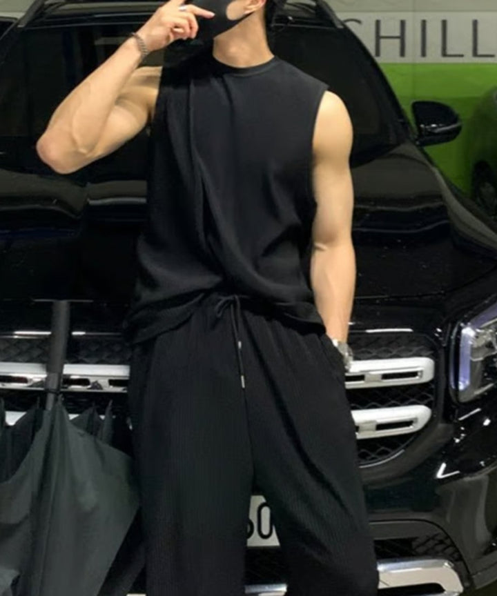 ブラックのプリーツノースリーブセットアップを着用し、黒い車の前でマスクに手を添えて佇む男性のスタイリングカット。