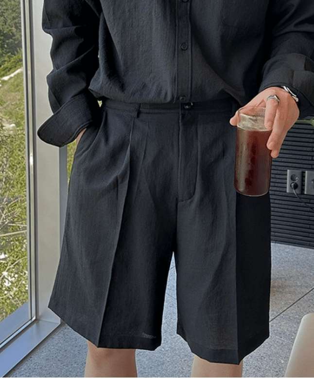 黒の長袖シャツをタックインし、フロントにタックが入った黒のショートパンツを穿いて、片手にアイスコーヒーを持つ男性のスタイリッシュなフロントカット。