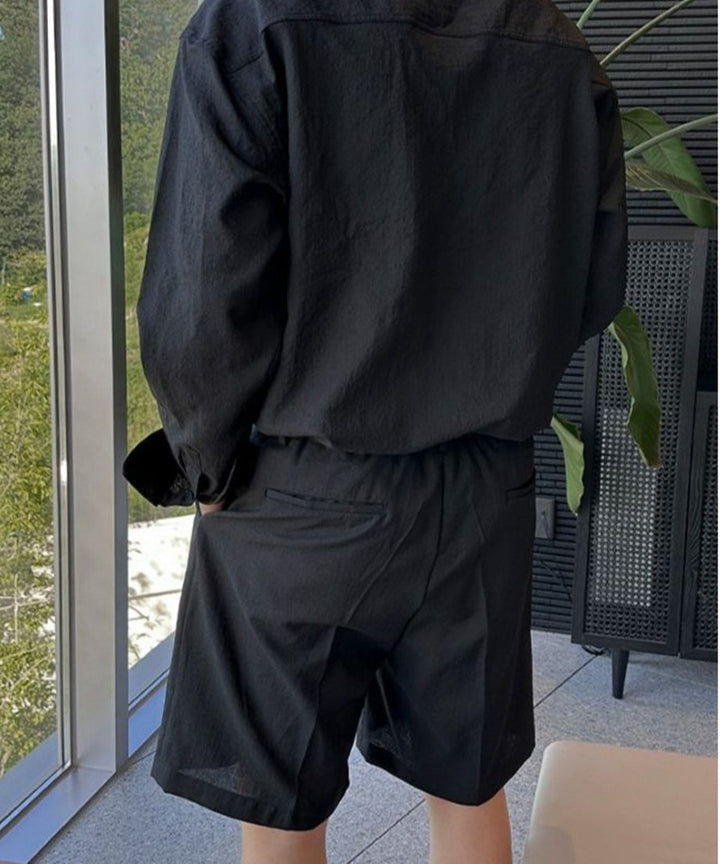 黒の長袖シャツに、後ろ身頃がすっきりとした黒のショートパンツを合わせ、バックポケットに手を入れながら窓際で佇む男性の後ろ姿。