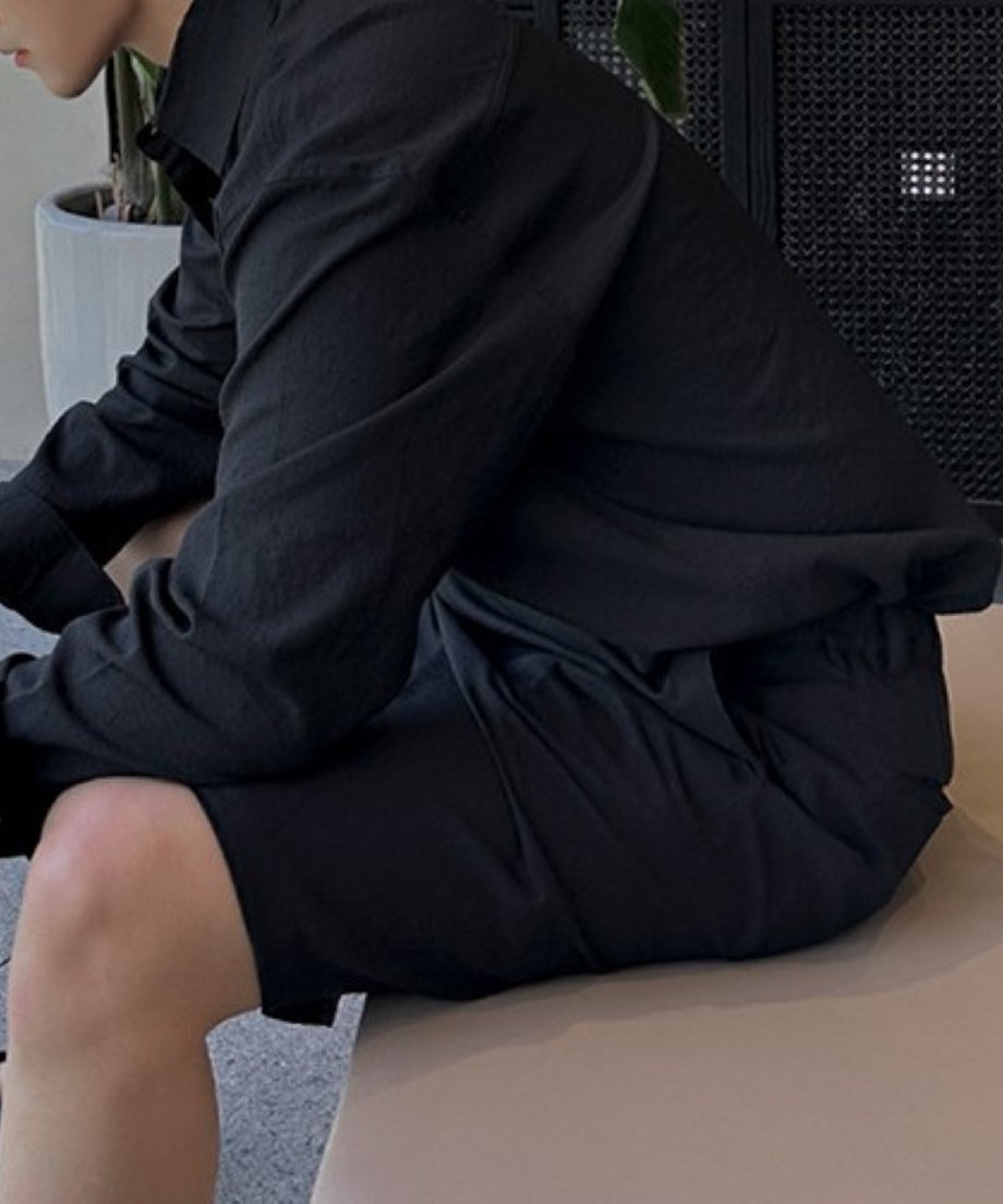 黒のシャツと黒のタックショートパンツを着用し、観葉植物のあるモダンな店内のベンチにリラックスして腰掛ける男性のサイドショット。