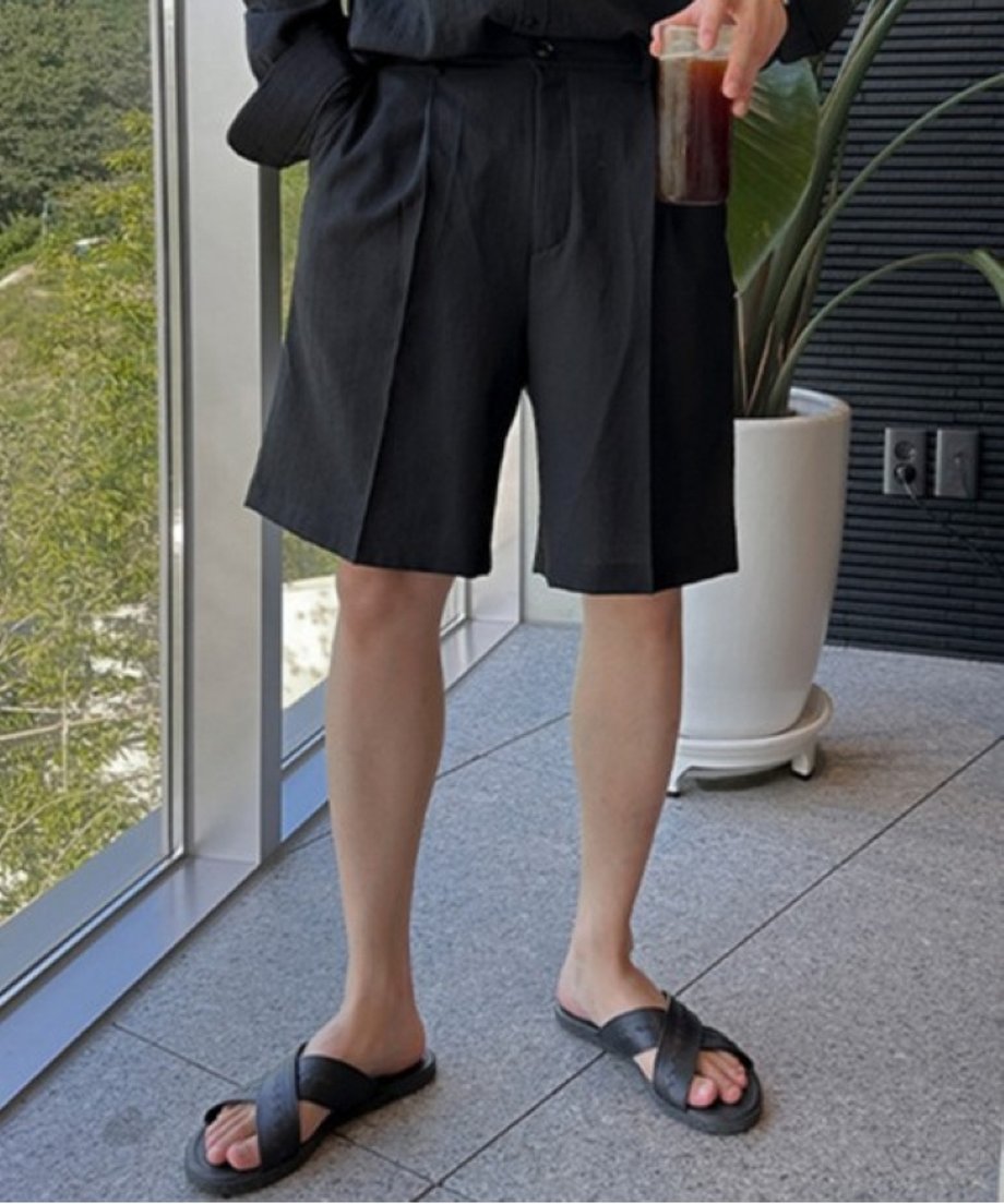 深めのフロントタックがきれいに映える黒のショートパンツに、黒のレザークロスサンダルを合わせた男性の、膝から下の足元クローズアップ。