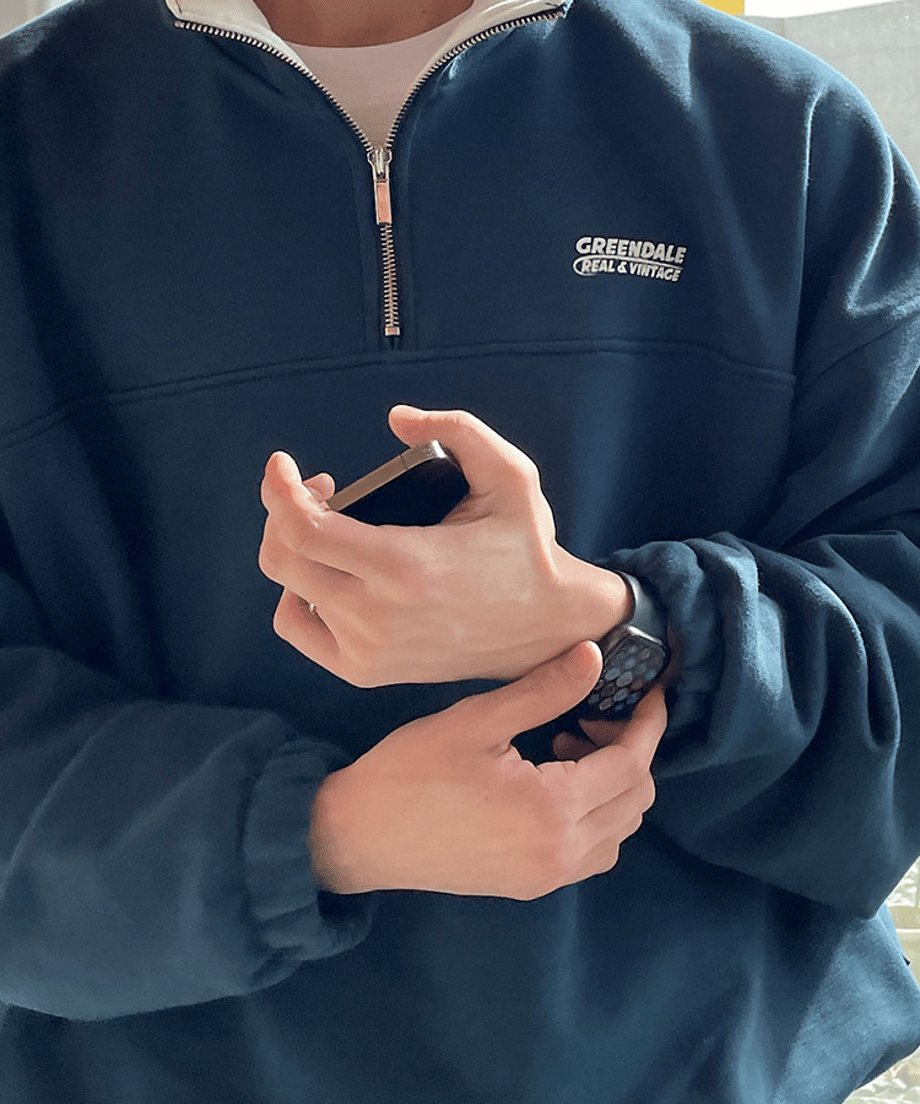プルオーバーを着用し、手元でスマートフォンを操作している男性のポーズカット。左胸にあしらわれた「GREENDALE REAL &amp; VINTAGE」のミニロゴ刺繍のサイズ感や、スマートなジッパーパーツの光沢感を捉えたビジュアル。