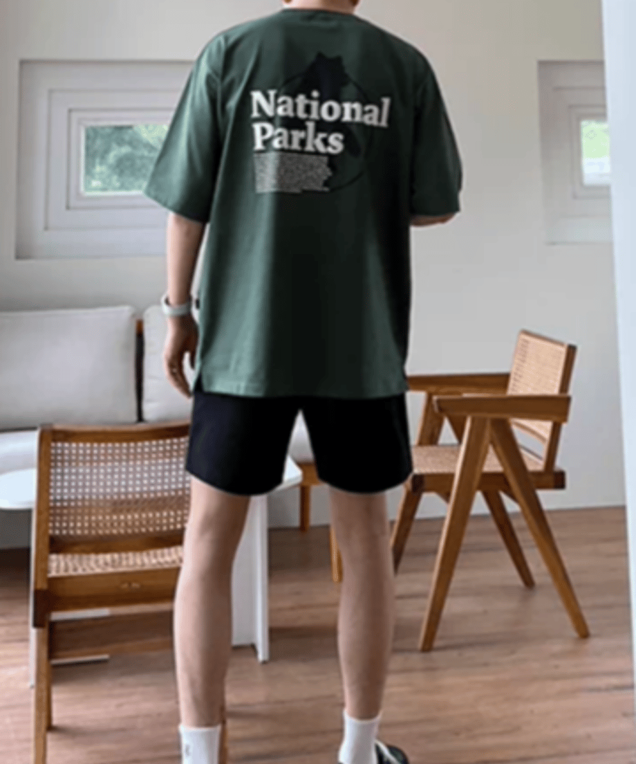 後ろ姿で直立する男性の全身スタイリング。オーバーサイズTシャツにブラックのハーフパンツ、白いソックスを合わせた、トレンド感のあるシティボーイ風のバックビュー。