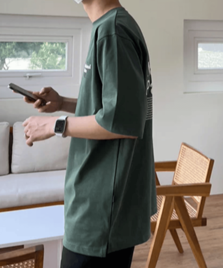 スマートフォンを片手に持ち、真横を向いて立つ男性のサイドビュー。裾のサイドスリットディテールと、前後の身頃が作り出す綺麗な直線的シルエットを強調した着用スナップ。