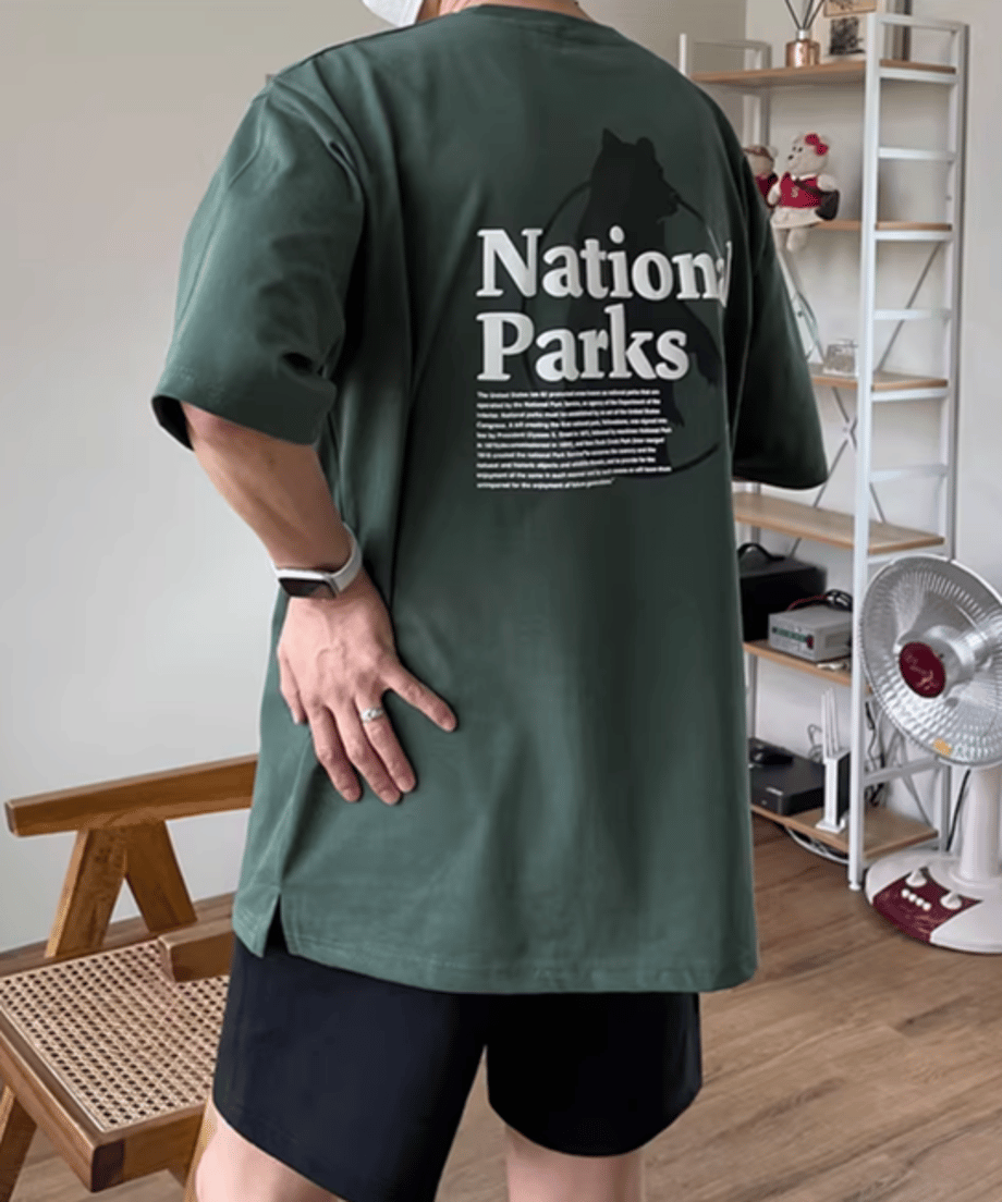 手を腰に当てて斜め後ろを振り返る男性のカット。背面に大胆にプリントされた「National Parks」の白いタイポグラフィと、その背景に佇むベア（熊）の黒いシルエットグラフィックが印象的な1枚。
