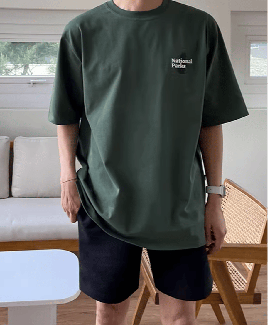 室内でダークグリーンのグラフィックTシャツの製品を着用した男性の半身ショット。ドロップショルダーのゆったりとしたシルエットと、左胸に施されたホワイトの「National Parks」ミニロゴデザインがクリアに伝わるフロントビュー。