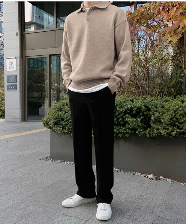 カーキのポロニットセーターと黒いスラックスを着用し、ポケットに手を入れて屋外に佇む人物の全身正面カット。
