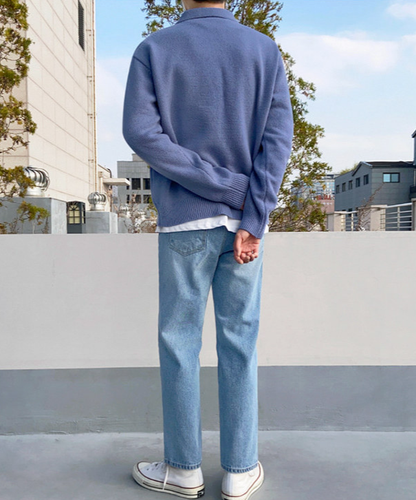 ブルーのポロカラー長袖ニットセーターにストレートデニムパンツ、白いスニーカーを合わせ、屋外に背を向けて立っている人物の後ろ姿。