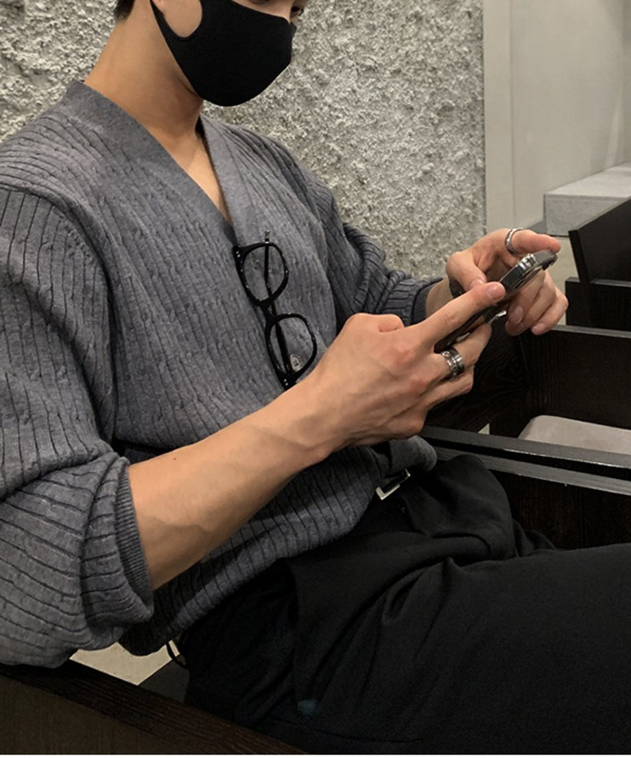 グレーのニットカーディガンを着用した男性が、カフェの椅子に腰掛けながらスマートフォンを操作している手元のアップカット。引き締まった前腕と、指先に光るシルバーリングが印象的な一枚。