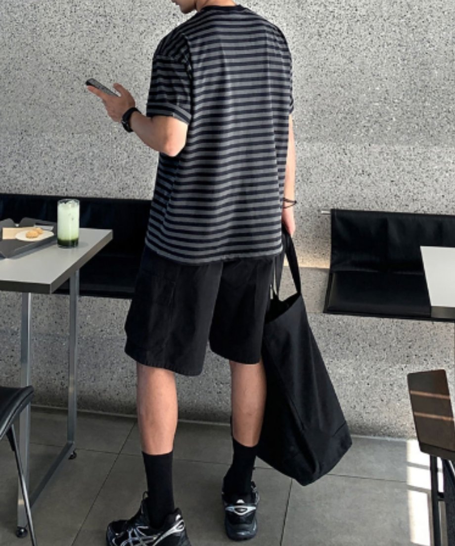 カフェテーブルの横に立ち、スマートフォンを見つめる男性の全身バック＆サイドスタイル。後ろから見たボーダーTシャツの綺麗なボックスシルエットと、ショーツを合わせた軽快なサマースタイル。
