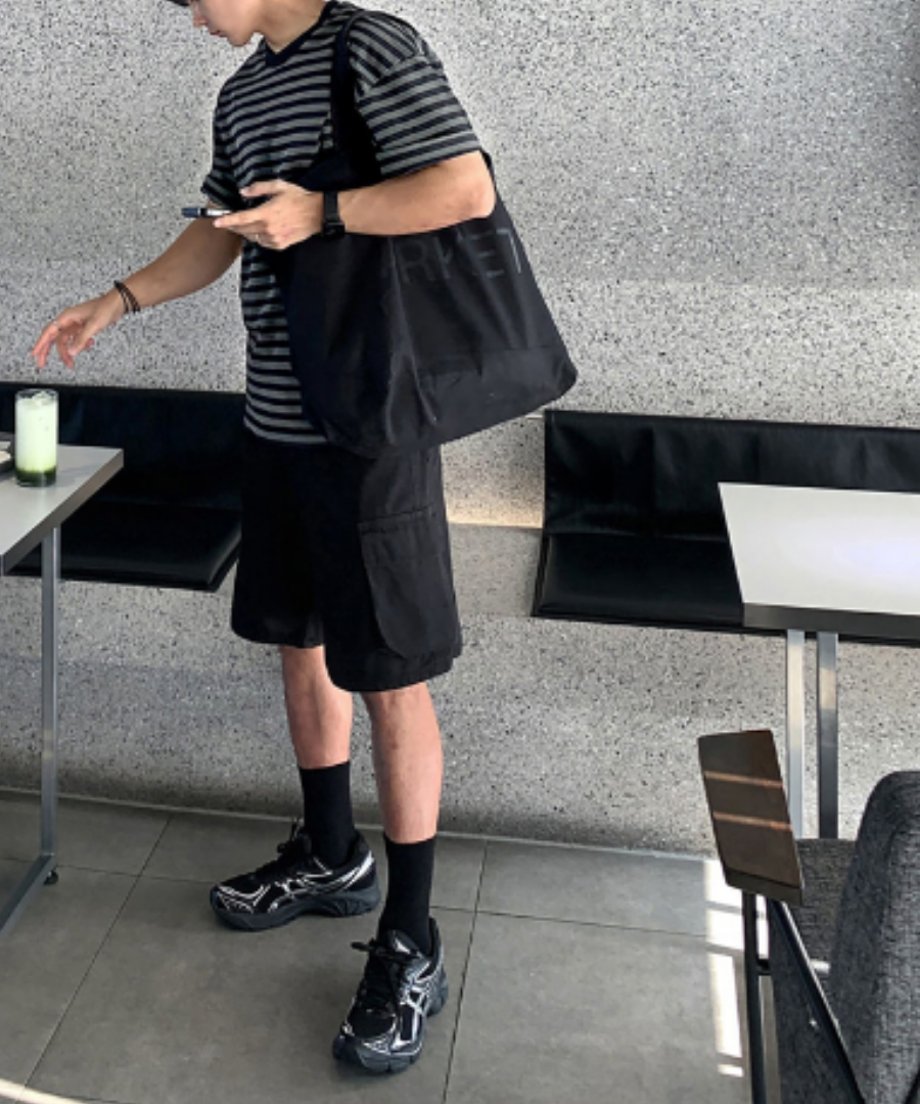 スタイリッシュなカフェの店内で、ボーダーTシャツに黒のカーゴショーツ、黒ソックスとハイテクスニーカーを合わせた男性の全身フロントスタイリング。左肩に大きな黒バッグを掛けたアーバンストリートなルック。