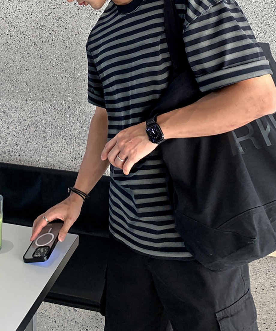 黒の大容量キャンバストートバッグを肩掛けし、白のカフェテーブルに手を置く男性のサイドスタイル。Tシャツのゆったりとした身幅の落ち感と、黒のボトムスを合わせたモノトーンコーデ。