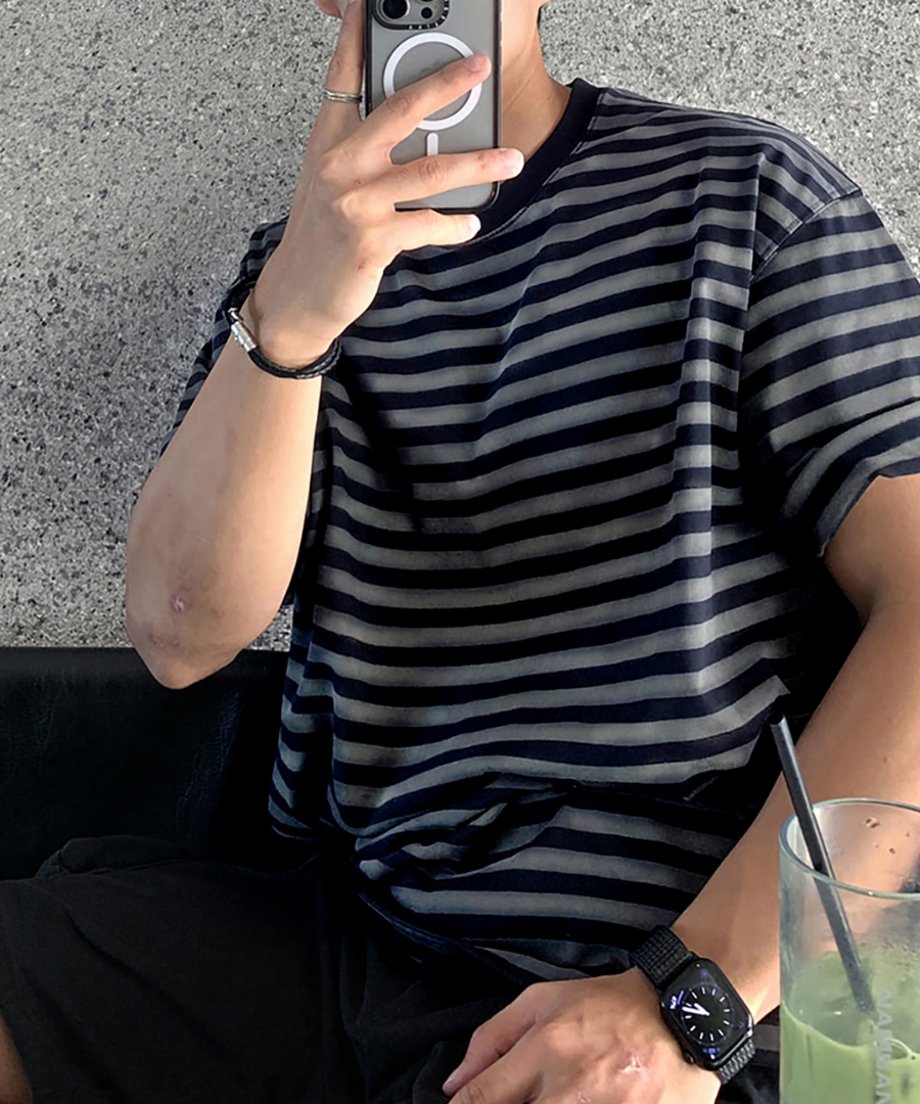 カフェの黒いベンチシートに腰掛け、スマートフォンで自撮り（セルフィー）をする男性のポーズカット。手前には緑色の抹茶ドリンクのグラスが置かれ、リラックスしたデイリールックを表現。