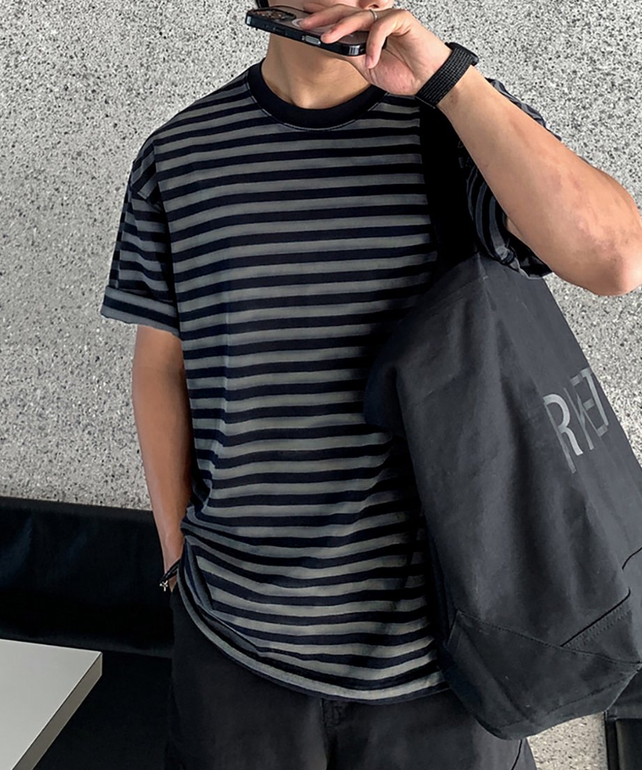 ウォッシュドボーダーTシャツを着用し、大きな黒のトートバッグを肩掛けした男性のフロントチェストアップ。片手でスマートフォンを持ち、袖口をロールアップしたストリートライクな佇まい。