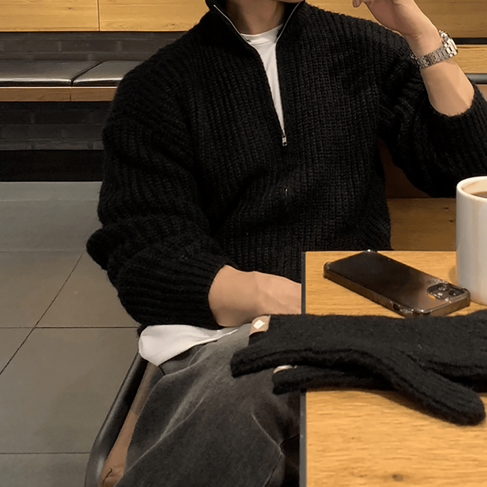 カフェのテーブル席で、顎に手を当てながらリラックスしている人物。ブラックのチャンキーリブハーフジップニットに白いTシャツの首元を覗かせ、シルバーの腕時計を合わせたシックなスタイリング。