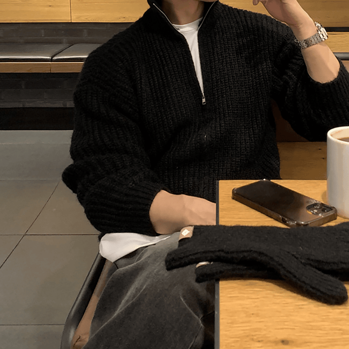 カフェのテーブル席で、顎に手を当てながらリラックスしている人物。ブラックのチャンキーリブハーフジップニットに白いTシャツの首元を覗かせ、シルバーの腕時計を合わせたシックなスタイリング。