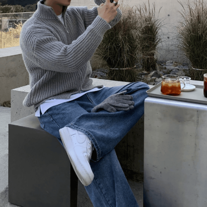 屋外のコンクリートベンチに腰掛け、脚を組んでスマートフォンを構える人物。グレーの厚手リブ編みハーフジップニットに、白Tシャツをレイヤードし、ウォッシュドブルーのワイドデニムと白いスニーカーを合わせたストリートカジュアルな着こなし。