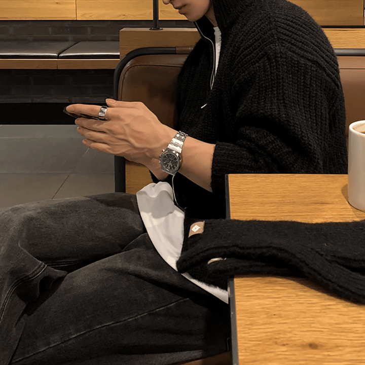 木製テーブルの横の席に座り、スマートフォンを操作する人物の手元のアップ。ブラックのチャンキーリブハーフジップニットを着用し、メタルバンドの腕時計とリングが上品なアクセントに。手前のテーブルには黒いニット手袋が置かれている。